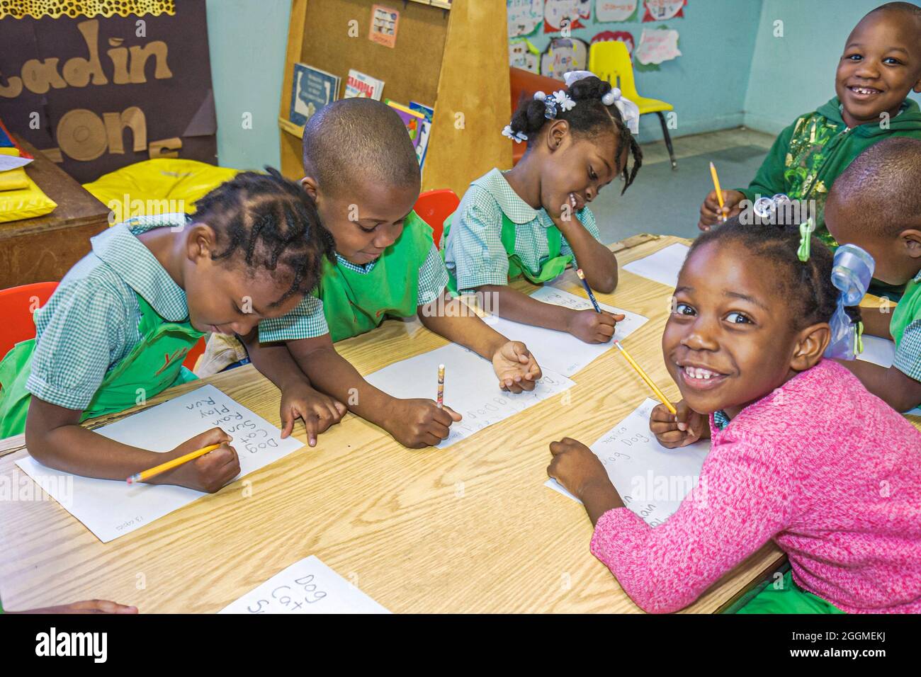 Young children in green school uniforms sitting around wooden table in colorful classroom, working on writing exercises with pencils and paper