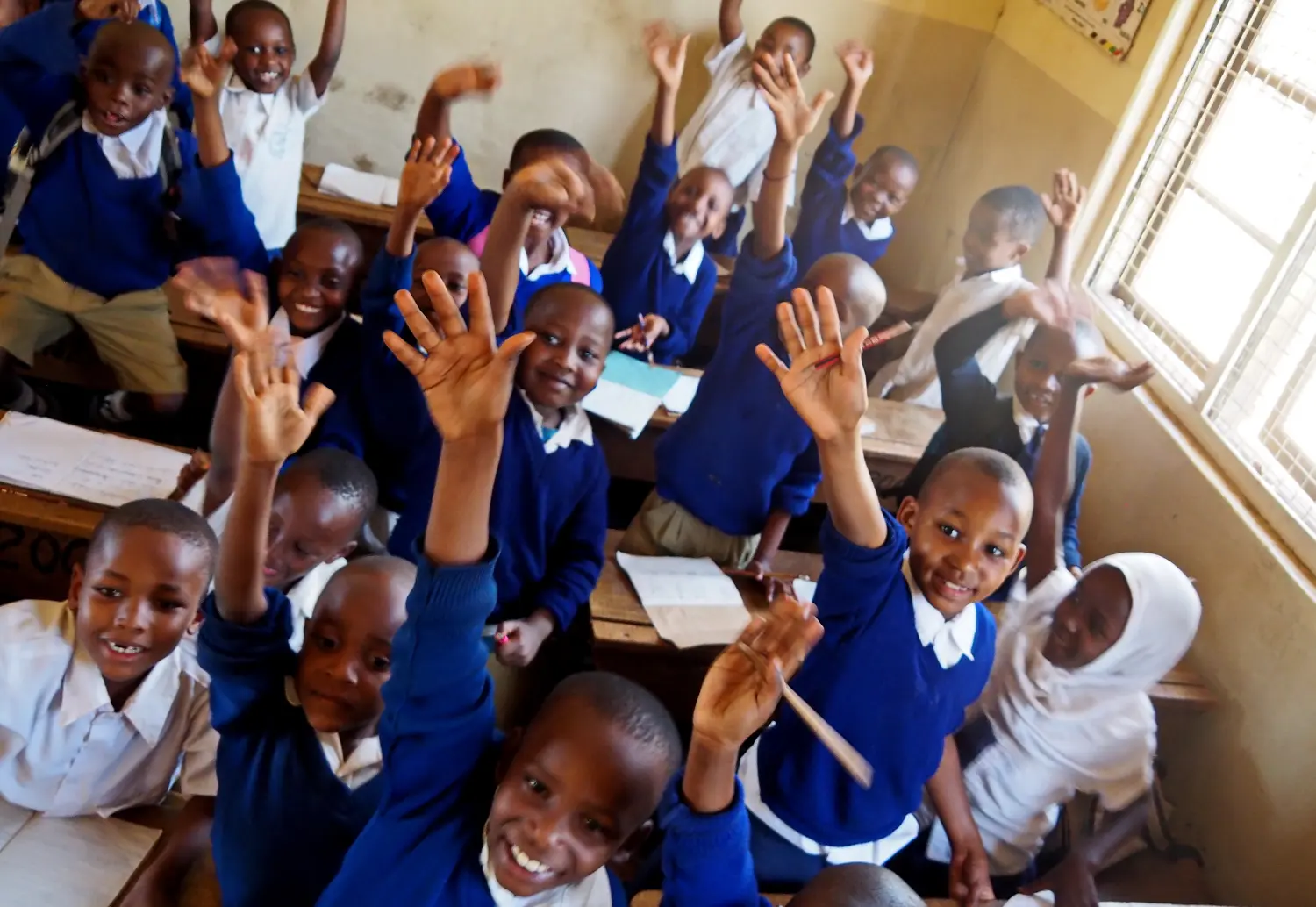 Enthusiastic students in blue and white school uniforms raising their hands and smiling in a sunlit classroom
