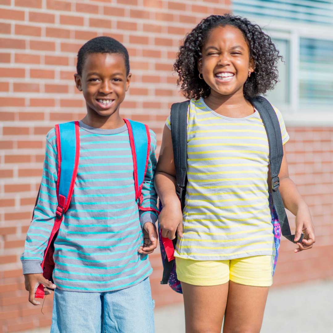 Two happy students with backpacks standing in front of brick school building - boy in blue striped shirt with red backpack and girl in yellow striped shirt with dark backpack